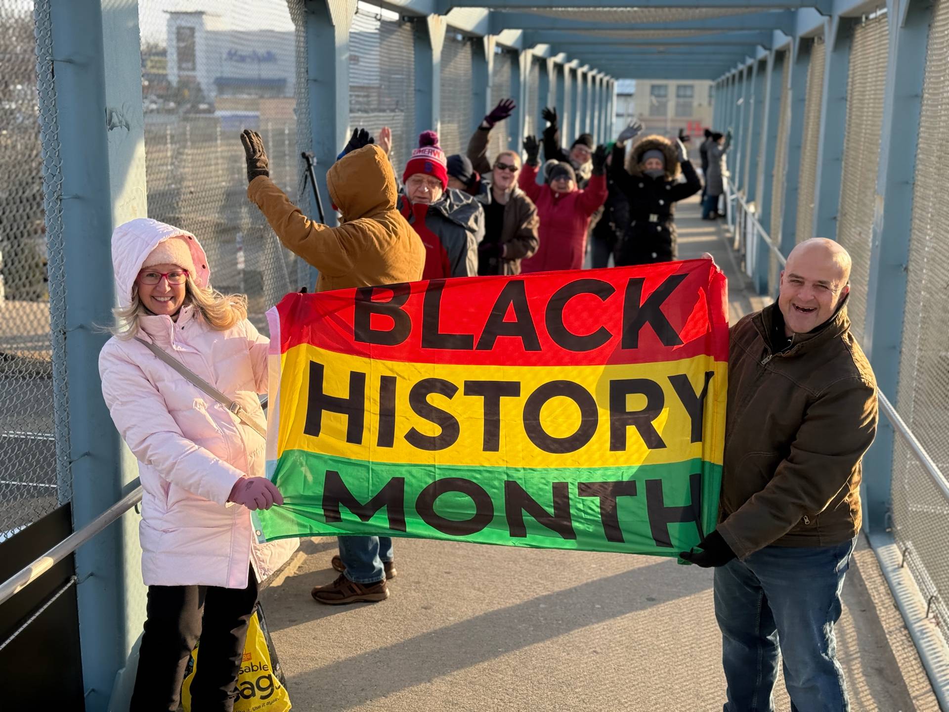 Protesters standing on a highway overpass holding a red, yellow, and green striped flag that says 'Black History Month'