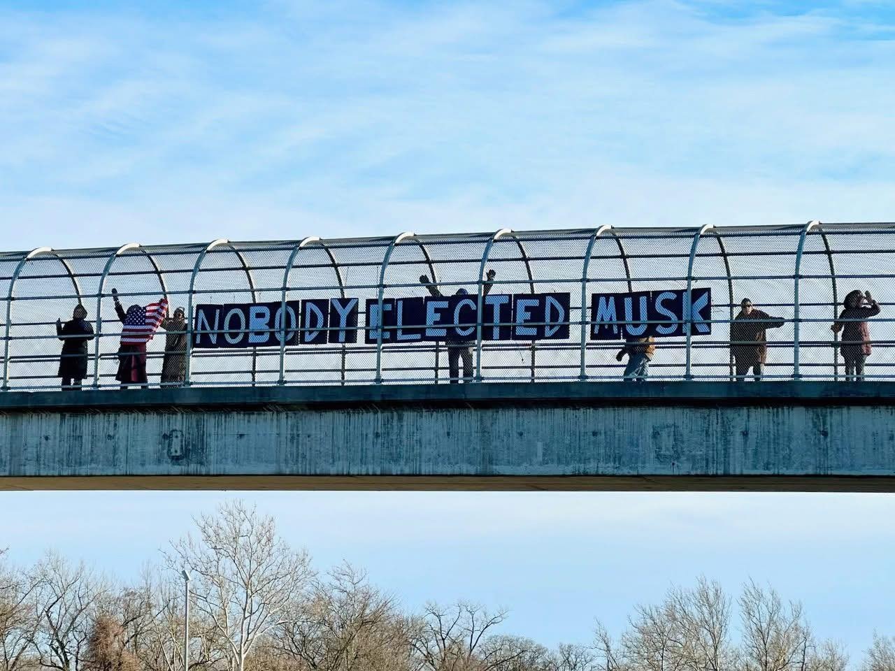 Protesters standing on a highway overpass holding a sign that says 'Nobody Elected Musk'