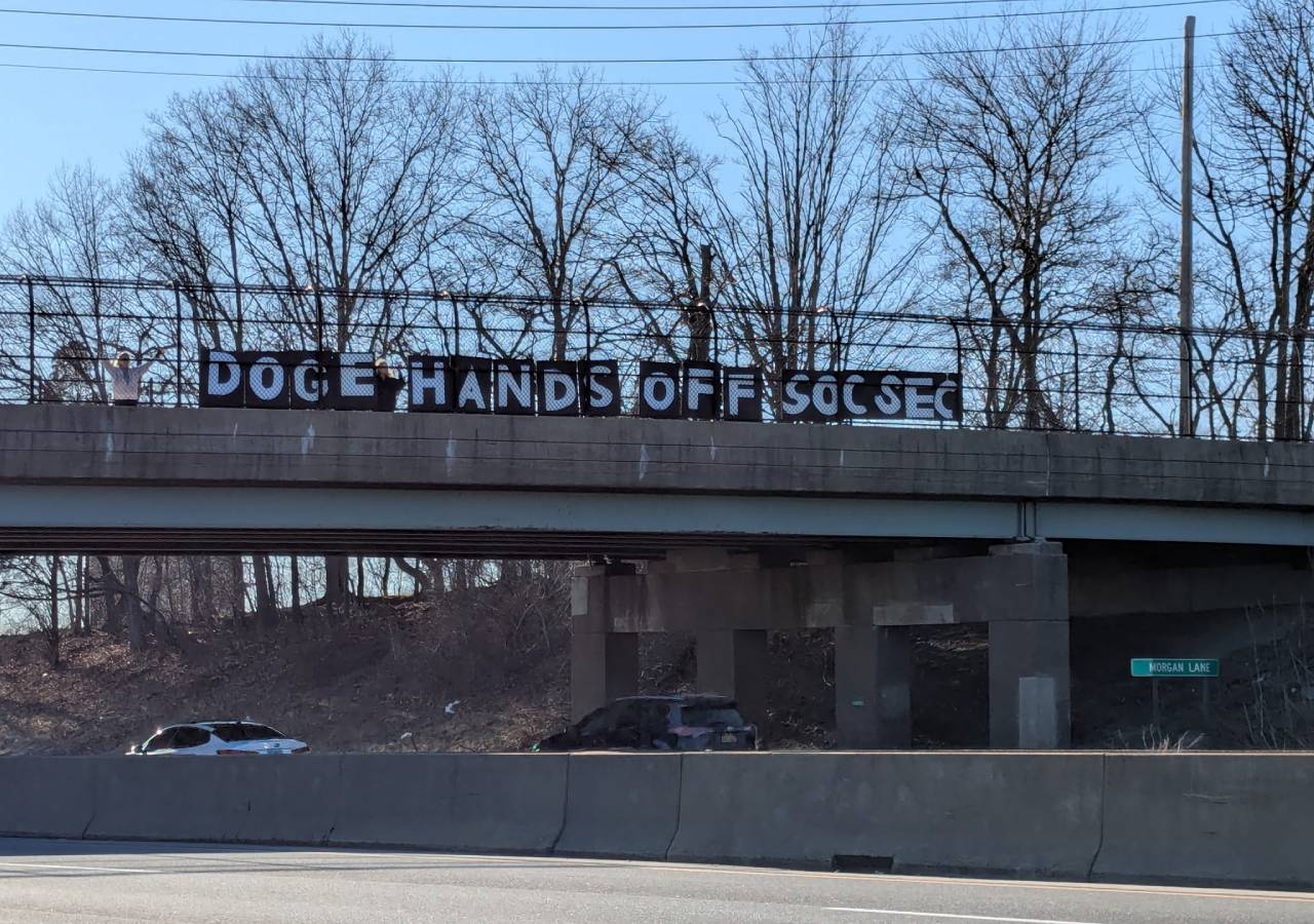 Protesters standing on a highway overpass holding a sign that says 'DOGE Hands Off Soc Sec'