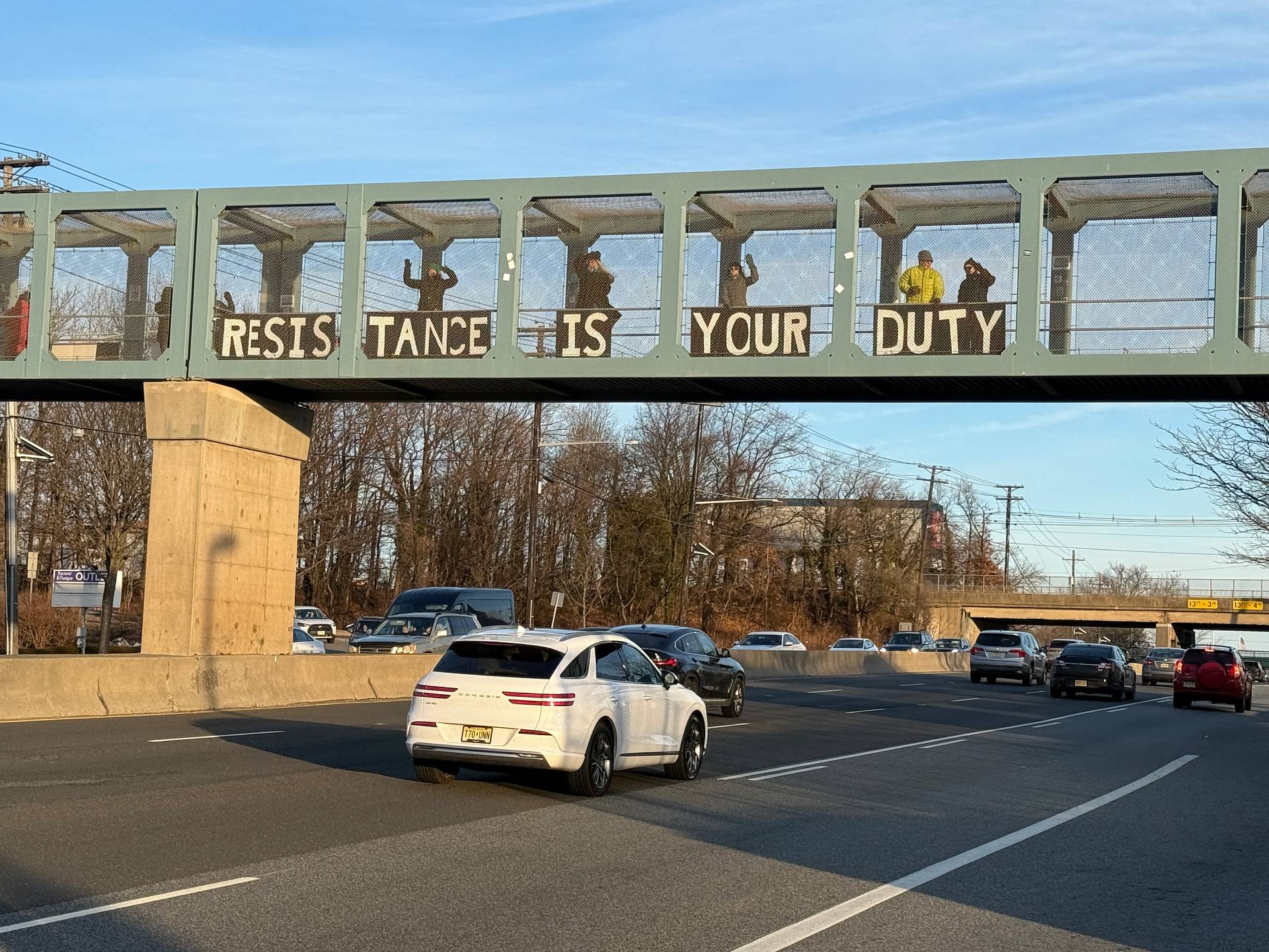 Protesters standing on a highway overpass holding a sign that says 'Resistance Is Your Duty'