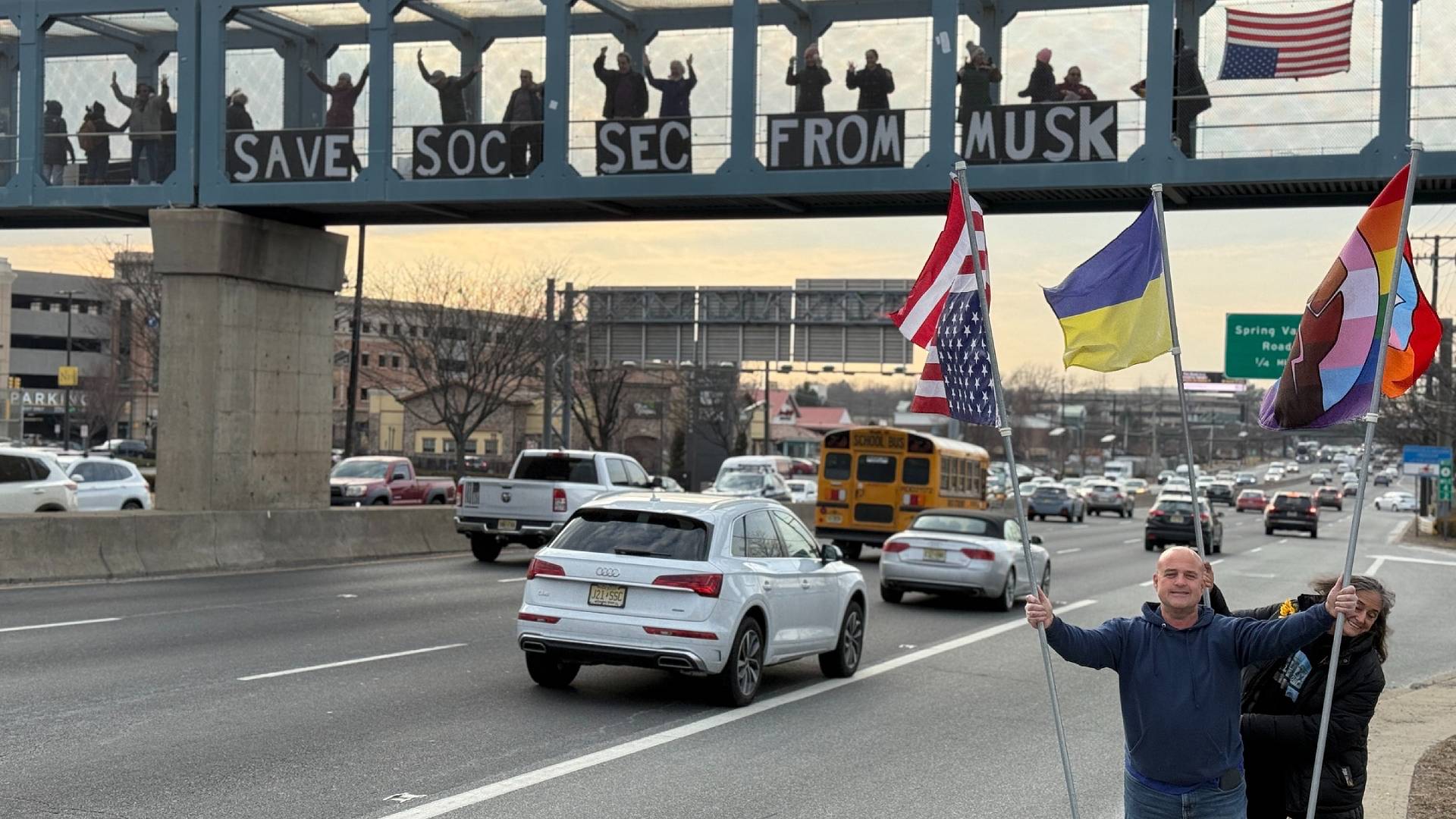 Protesters standing on a highway overpass holding a sign that says 'Save Soc Sec From Musk'