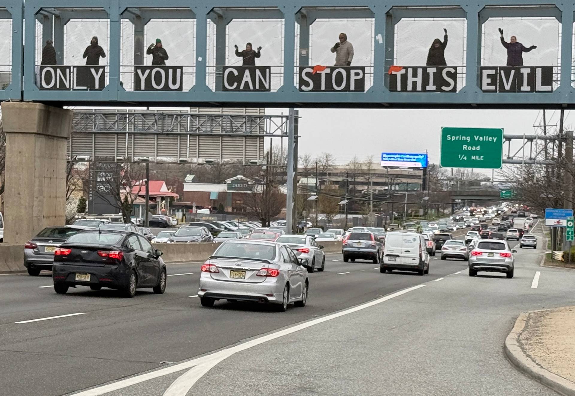 Protesters standing on a highway overpass holding a sign that says 'Only You Can Stop This Evil'