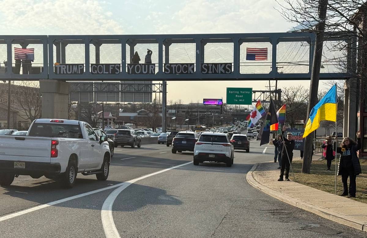 Protesters standing on a highway overpass holding a sign that says 'Trump Golfs Your Stock Sinks'